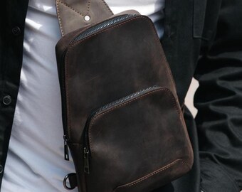 May include: A dark brown leather sling bag featuring a vertical design with a main zippered compartment and a smaller front zippered pocket. The bag includes a single adjustable shoulder strap attached with a metal ring and dark metal hardware.