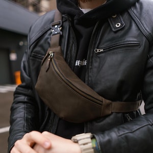 Può includere: Un marsupio in pelle marrone scuro con finitura opaca, chiusura a zip curva con tiretti in pelle e una fibbia tattica in metallo nero sulla tracolla regolabile. La borsa è indossata a tracolla sopra una giacca di pelle nera.