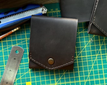 May include: A dark brown leather wallet with a single brass snap closure and visible stitching along the flap. The wallet is positioned on a green cutting mat next to a metal ruler that shows measurements in inches and centimeters.