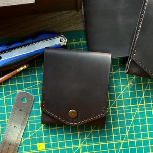 May include: A dark brown leather wallet with a snap closure. The wallet is square-shaped and has a stitched border. A metal ruler and a utility knife are visible in the background.