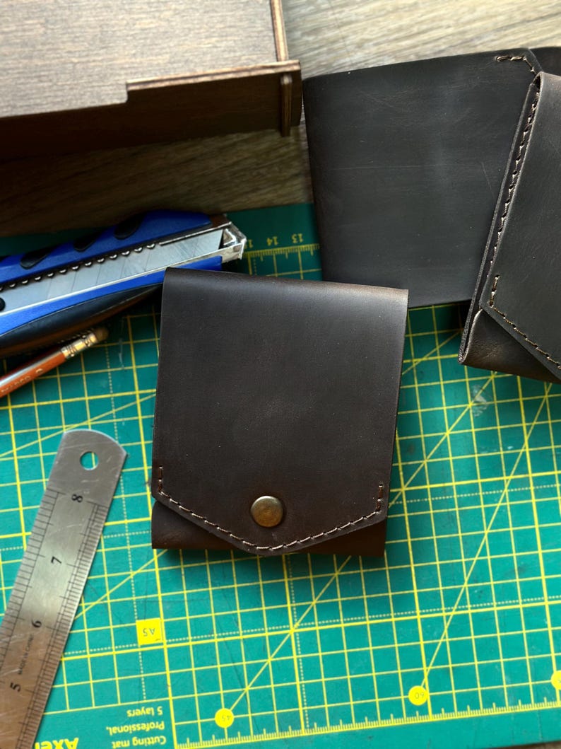 May include: A dark brown leather wallet with a snap closure. The wallet is square-shaped and has a stitched border. A metal ruler and a utility knife are visible in the background.