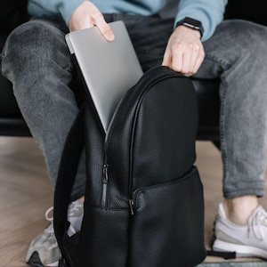 May include: A black textured leather backpack with a main zippered compartment and a front pocket. The backpack features silver-toned metal zippers and is shown with a silver laptop being placed into the main compartment of the bag.