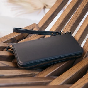 May include: A rectangular black leather wallet featuring a wrap-around zipper closure and a detachable wrist strap. The wallet has a smooth, matte finish and is constructed with visible stitching along the edges, including a metal clasp attachment.