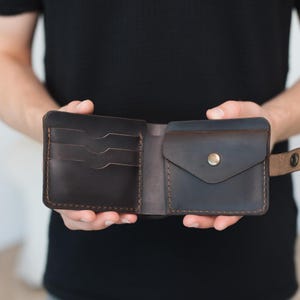 May include: A dark brown leather bifold wallet featuring three horizontal card slots on the left side and a coin pocket with a brass-colored snap button closure on the right side. The wallet is constructed with visible brown stitching throughout.
