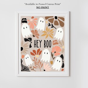 May include: A framed print featuring a beige background with white ghost illustrations and floral designs in shades of brown, peach, and yellow. The phrase "HEY BOO" is written in black. The print is in a white frame.