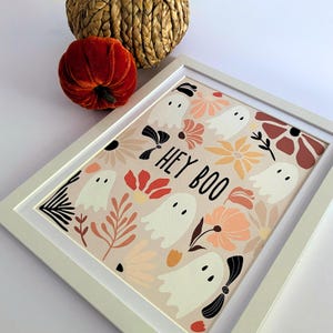 May include: A framed print with a Halloween theme. The artwork features white ghost illustrations among floral designs in shades of peach, orange, and brown. The phrase "HEY BOO" is printed in black. A small orange velvet pumpkin and a woven basket are in the background.