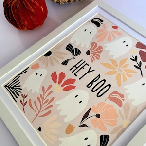 May include: A framed print featuring a pattern of white ghosts and floral designs in shades of peach, orange, and black on a light pink background. The words "HEY BOO" are printed in black. The frame is white.