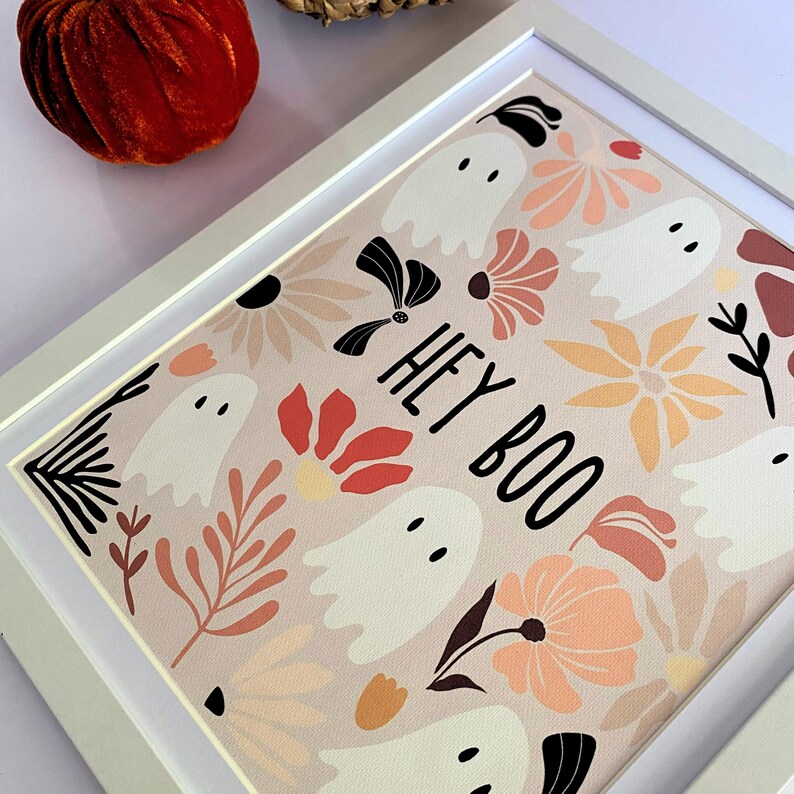 May include: A framed print featuring a pattern of white ghosts and floral designs in shades of peach, orange, and black on a light pink background. The words "HEY BOO" are printed in black. The frame is white.