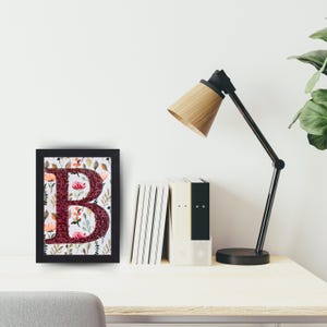 May include: A framed print of the letter "B" with a floral design. The letter is made of burgundy fabric and the flowers are in shades of pink, orange, and green. The frame is black.