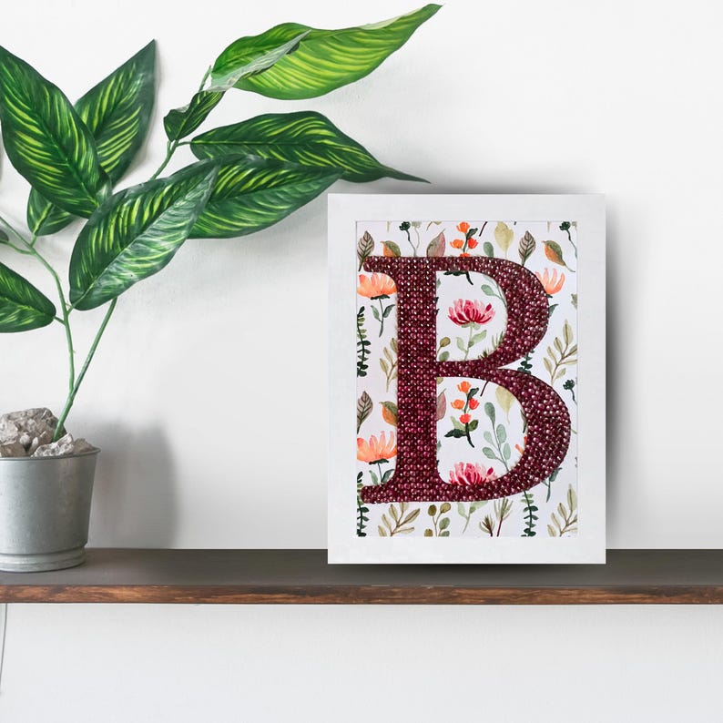 May include: A framed letter B with a burgundy colored background. The letter is made of small, sparkly beads. The background is a white floral pattern with pink and orange flowers.