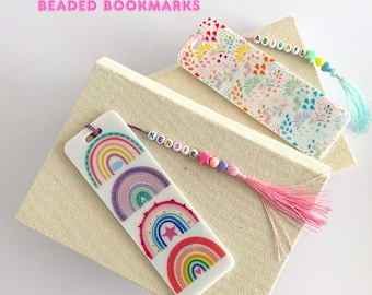 May include: Two colorful beaded bookmarks with tassels. One bookmark features a rainbow design with a star and the name "KENSIE" spelled out in beads. The other bookmark features a floral design with the name "ABIGAIL" spelled out in beads.