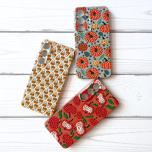 May include: Three rectangular phone cases featuring raised dot patterns. One case has a cream background with candy corn, one is grey with orange pumpkins and green leaves, and one is terracotta red with red and white apple slices and green leaves.