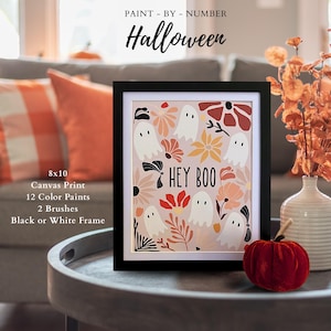 May include: A framed Halloween-themed paint-by-number canvas print. The artwork features white ghosts, flowers, and the phrase "HEY BOO" on a light pink background. The frame is black. The canvas print measures 8x10 inches.