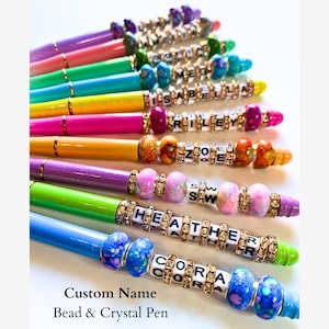 May include: A set of colorful pens with custom names spelled out in beads and crystals. The pens are arranged in a rainbow pattern, with the names "CORA", "HEATHER", "SW", "ZOE", "RILEY", "ISABELLA", "MEG", and others visible.