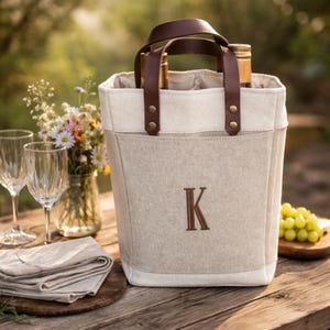 May include: A beige wine tote bag with a brown leather handle and the letter "K" embroidered on the front. The bag holds two wine bottles. The background includes wine glasses, flowers, and grapes.