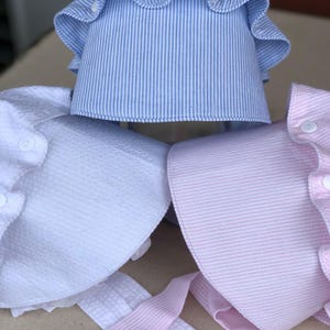 May include: Three baby bonnets in blue and white striped, white textured, and pink and white striped fabrics. Each bonnet has a ruffled brim and is secured with buttons. Pink and white striped ties are visible.
