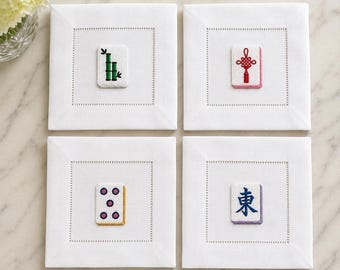 May include: A set of four square white cloth napkins, each featuring a centered embroidered mahjong tile design. The designs include a green bamboo stalk, a red Chinese knot, five purple dots, and a blue East character on a light purple tile.