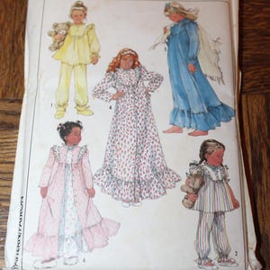 May include: A vintage Simplicity sewing pattern envelope featuring illustrations of children's sleepwear. The designs include pajamas, nightgowns, and robes in various styles and colors. The envelope is slightly worn, with the Simplicity logo prominently displayed.