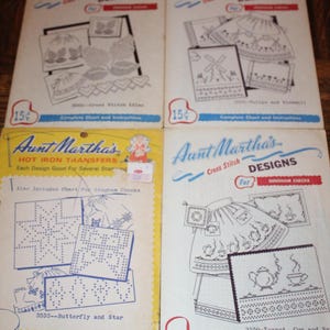 May include: Four vintage Aunt Martha's pattern packets for cross stitch and hot iron transfers. The white and yellow paper envelopes feature black line art illustrations of various designs including flowers, windmills, teapots, and stars.