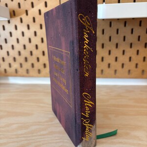 May include: A copy of "Frankenstein" by Mary Shelley. The book features a dark purple cover with gold lettering and a quote. A green ribbon bookmark is visible.
