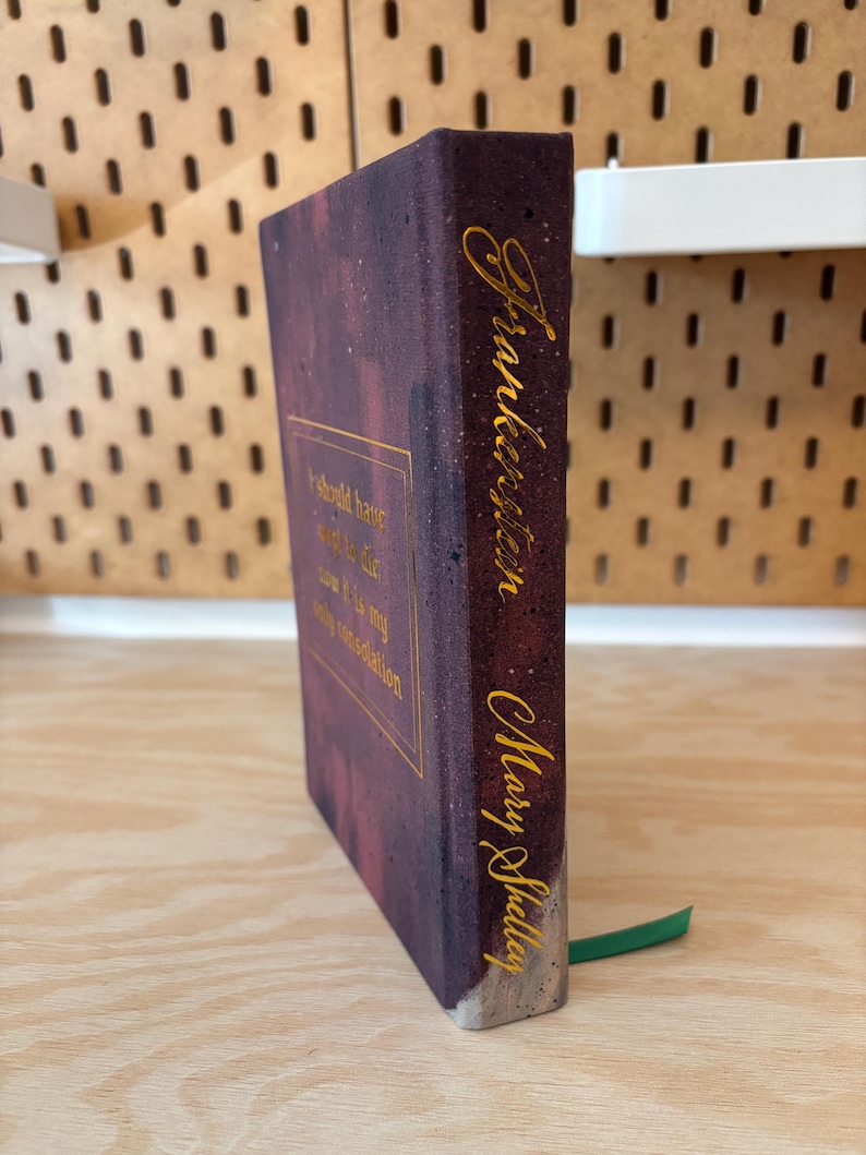 May include: A copy of "Frankenstein" by Mary Shelley. The book features a dark purple cover with gold lettering and a quote. A green ribbon bookmark is visible.
