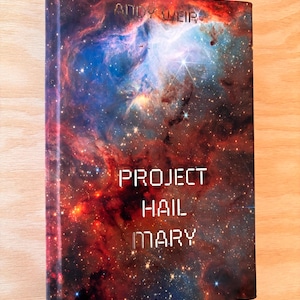 May include: A hardcover book titled "Project Hail Mary" by Andy Weir. The cover features a vibrant image of a nebula with red, blue, and white colors. The title is in a white, pixelated font.