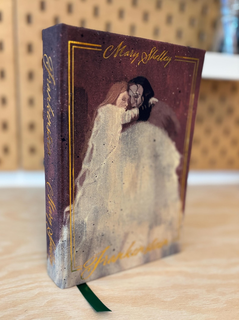 May include: A burgundy hardcover book with gold lettering and an illustration of two figures. The book's title, "Frankenstein," and author, Mary Shelley, are displayed. A green ribbon bookmark is visible.