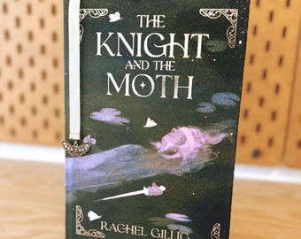Puede incluir: Un libro de tapa dura titulado The Knight and the Moth de Rachel Gillig con una cubierta verde oscuro que muestra una figura etérea púrpura y polillas blancas. Un marcapáginas de cinta blanca con un pequeño dije de mariposa de metal.