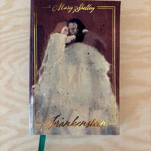 May include: A hardback book with a burgundy cover. Gold lettering spells out "Mary Shelley" and "Frankenstein." The cover art is a painting of two figures embracing. A green ribbon bookmark is present.