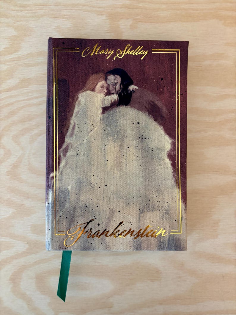 May include: A hardback book with a burgundy cover. Gold lettering spells out "Mary Shelley" and "Frankenstein." The cover art is a painting of two figures embracing. A green ribbon bookmark is present.