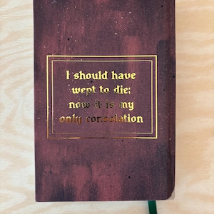 May include: A burgundy hardback book with gold lettering. The text within a gold rectangle reads, "I should have wept to die; now it is my only consolation." A green ribbon bookmark is visible.