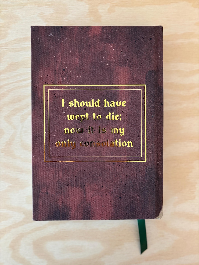 May include: A burgundy hardback book with gold lettering. The text within a gold rectangle reads, "I should have wept to die; now it is my only consolation." A green ribbon bookmark is visible.