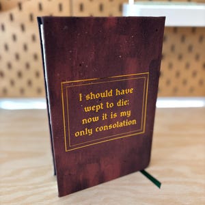 May include: A maroon journal with gold lettering that reads "I should have wept to die; now it is my only consolation." The journal has a green ribbon bookmark. The cover has a textured, distressed appearance.