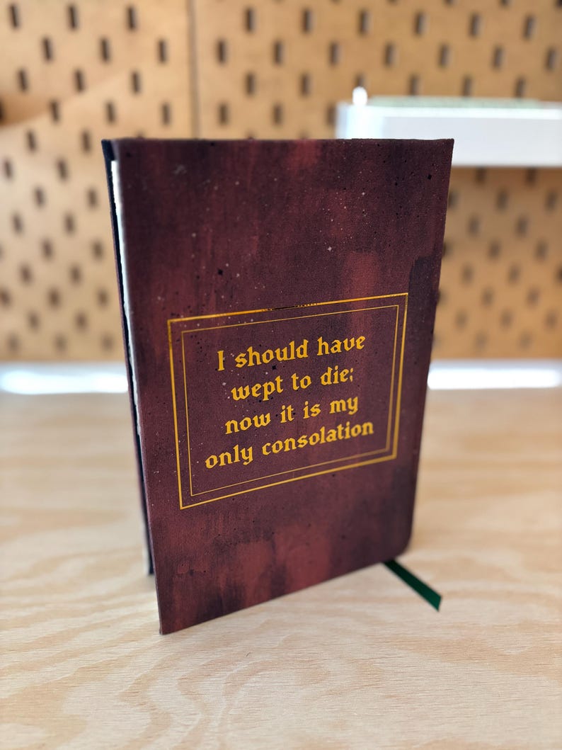 May include: A maroon journal with gold lettering that reads "I should have wept to die; now it is my only consolation." The journal has a green ribbon bookmark. The cover has a textured, distressed appearance.