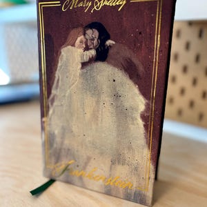 May include: A hardback book featuring a painting of a couple embracing. The title "Frankenstein" is in gold script, with "Mary Shelley" above. A green ribbon bookmark is visible. The book is on a wooden surface.
