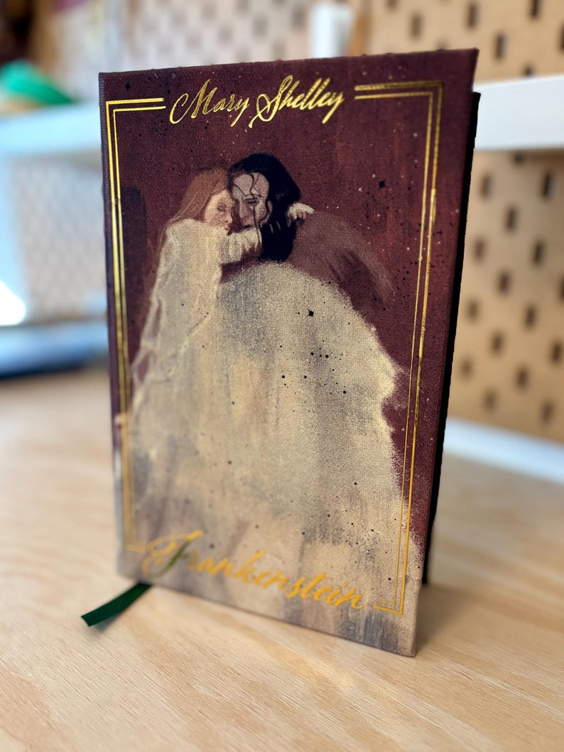 May include: A hardback book featuring a painting of a couple embracing. The title "Frankenstein" is in gold script, with "Mary Shelley" above. A green ribbon bookmark is visible. The book is on a wooden surface.