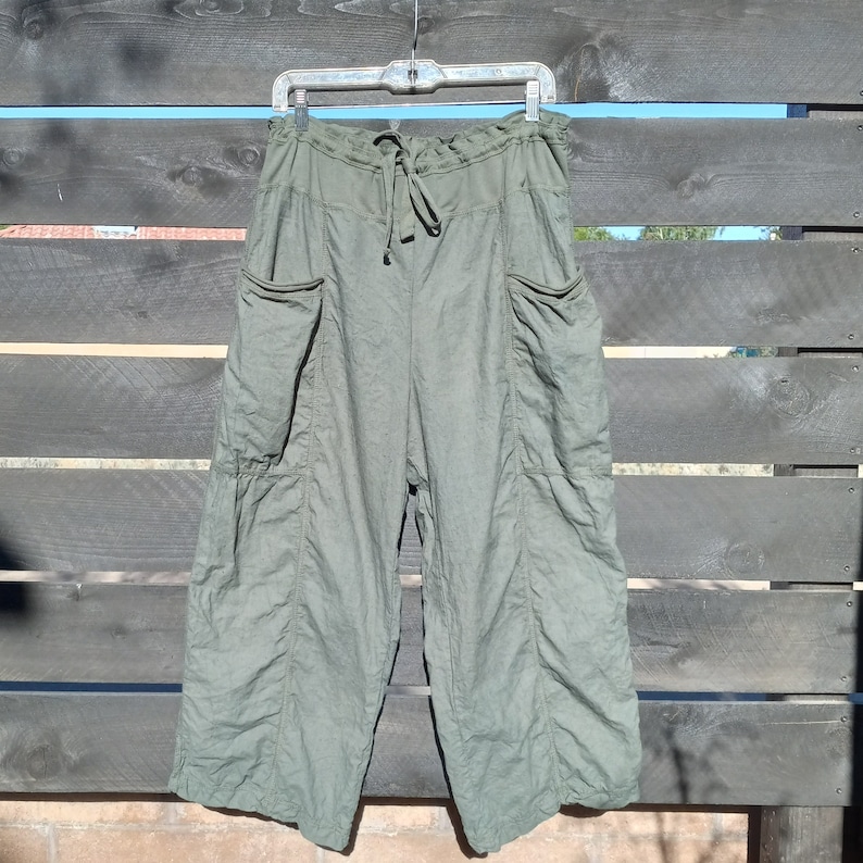 Puede incluir: Pantal&oacute;n de lino verde con cintura de cord&oacute;n y bolsillos grandes. El pantal&oacute;n est&aacute; colgado en una percha contra un fondo de madera.