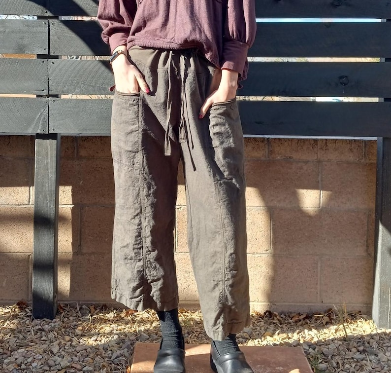 May include: A pair of brown linen trousers with a wide leg and a drawstring waist. The trousers have two large pockets on the front and are worn with black shoes.