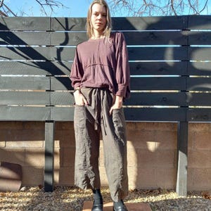 May include: A woman wearing a loose-fitting, long-sleeved, mauve shirt with brown, wide-legged, cropped trousers. The trousers have large pockets on the front.