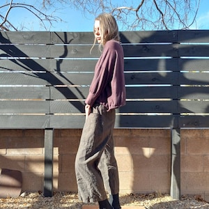 May include: A person wearing a maroon, long-sleeved shirt with a loose fit and brown, wide-leg trousers. The person is standing in front of a black wooden fence.