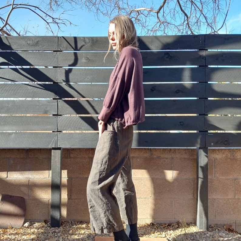 May include: A person wearing a maroon, long-sleeved shirt with a loose fit and brown, wide-leg trousers. The person is standing in front of a black wooden fence.