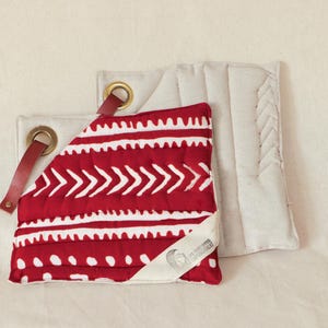 Può includere: Due presine rosse e bianche con motivi, con retro in tessuto bianco e anelli in pelle marrone. Le presine hanno un'etichetta bianca con il testo "Quilted" su di esse.