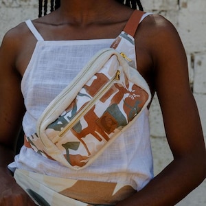 Può includere: Marsupio in tela con motivo mimetico marrone, bianco e verde. La borsa ha una cerniera dorata e un cinturino marrone. La borsa è indossata a tracolla. La modella indossa una canotta bianca.