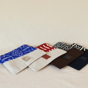 May include: Five fabric pouches with different patterns. The pouches are folded on top of each other. The top pouch is blue with a white geometric pattern. The second pouch is red with a white geometric pattern. The third pouch is black and white with a geometric pattern. The fourth pouch is brown with a black flap. The fifth pouch is navy blue with a black flap.