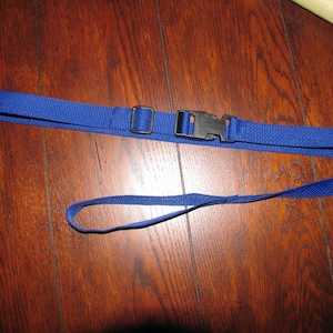 May include: A blue nylon strap with a black plastic buckle and a metal square buckle. A smaller blue nylon strap is looped at one end. The straps are lying on a wooden surface.