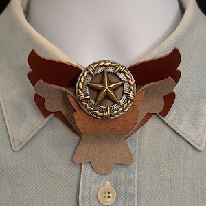 May include: A brown leather bow tie with a star-shaped metal embellishment in the center. The bow tie is on a light blue collared shirt.