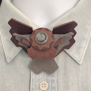 May include: A brown, gray, and purple leather bow tie with a circular, gray, and brown button in the center.