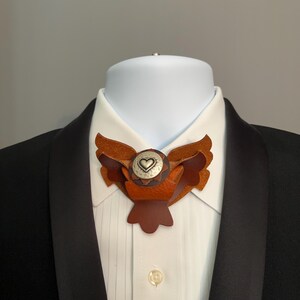 May include: A brown leather bow tie with a layered wing design and a silver-toned center featuring a heart. The bow tie is worn on a white collared shirt under a black suit jacket, suitable for formal events.