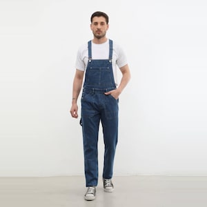 Puede incluir: Un peto de mezclilla azul con un bolsillo en el peto, tirantes ajustables con botones de metal y dos bolsillos delanteros, usado sobre una camiseta blanca de manga corta con cuello redondo y zapatillas de lona blancas.