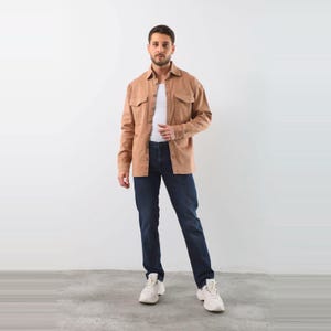 Könnte beinhalten: Ein beiges, langärmeliges Hemd mit Knopfleiste über einem schlichten weißen T-Shirt, kombiniert mit einer dunkelblauen Jeans und weißen Turnschuhen. Das Hemd hat zwei Brusttaschen mit Klappen und einen klassischen Kragen für einen Lagenlook.